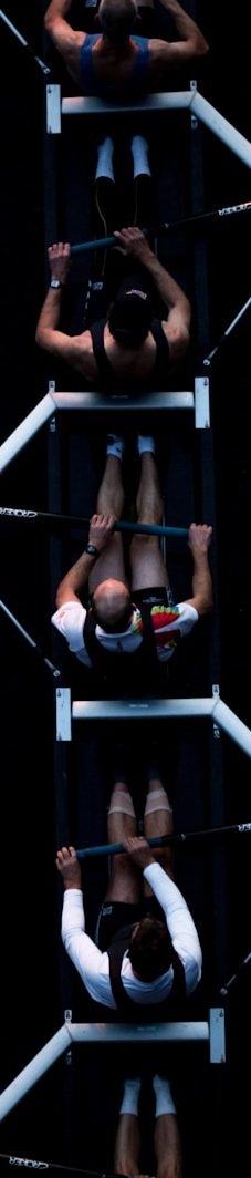 Vier Personen trainieren in Rudergeräten, mit unterschiedlicher Körperhaltung.